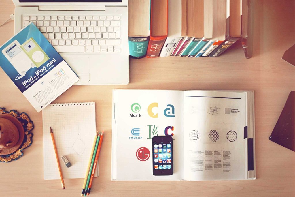 Buch mit Markenlogos liegt auf einem Schreibtisch mit Laptop, Büchern und Schreibutensilien sowie einem iPhone, welches auf dem Buch liegt